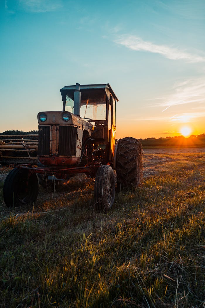 An old tractor sits in a rural field during a vibrant sunrise, symbolizing agriculture.